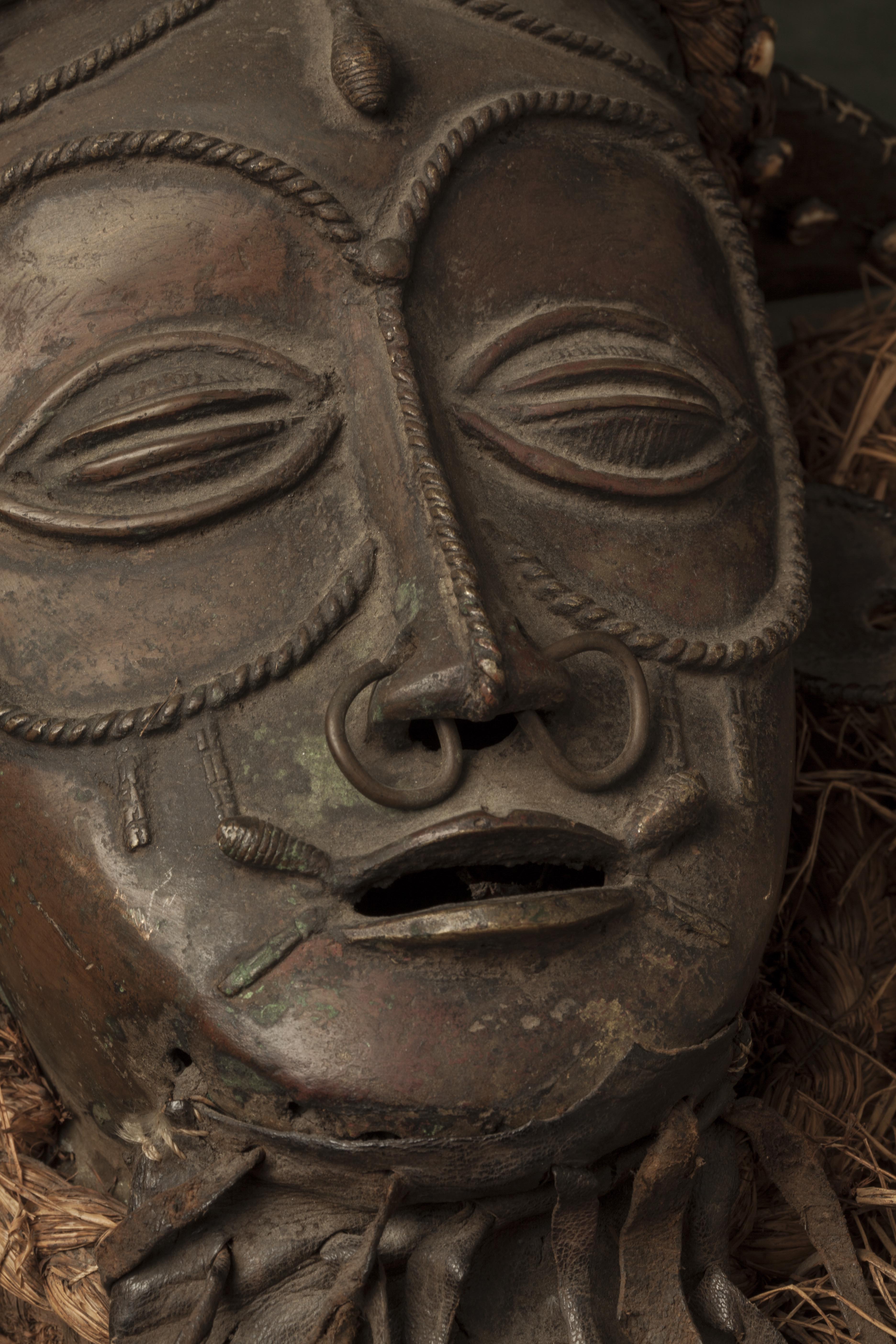 Br.  Benin (masque), d`afrique : Nigéria, statuette Br.  Benin (masque), masque ancien africain Br.  Benin (masque), art du Nigéria - Art Africain, collection privées Belgique. Statue africaine de la tribu des Br.  Benin (masque), provenant du Nigéria, 108/680.Très ancien masque en bronze h.27cm.
Les yeux mi-clos,entouré de scarifications. En plus des scarifications sur le front et le nez.Il porte de anneaux dans le nez et a des oreilles rondes  trouées.Il porte une barbe en cuir.La coiffure est faite d